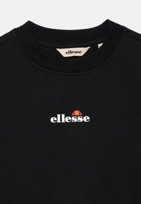 Melnas kreklu ar apaļu kaklu, ar baltu 'ellesse' logotipu un oranžu pusapļa dizainu centrā uz krūtīm, kā arī atbilstošu etiķeti iekšējā apkaklē.
