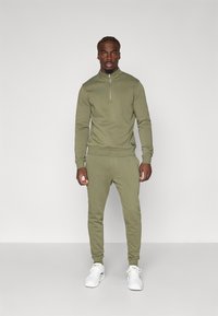 Felpa con zip di colore verde oliva e pantaloni jogger coordinati in materiale morbido. Accentuati da un piccolo logo sull'orlo, abbinati a scarpe da ginnastica bianche.