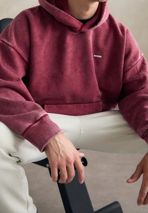 Man draagt een vervaagde rode hoodie en lichtbeige broek, zittend met handen rustend op zijn knieën tegen een neutrale achtergrond.