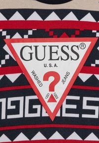 Rød og sort mønstret stof med en trekantet hvid etikette. Etiketten har "GUESS®," "WASHED JEANS," og et rødt spørgsmålstegn design.