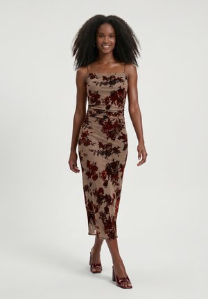 Femme aux cheveux bouclés marchant dans une robe midi beige sans manches avec un motif floral rouge foncé et des sandales à talons hauts bordeaux.