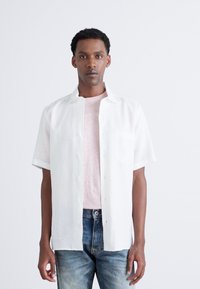 Camisa blanca de manga corta, con botones y bolsillo, llevada sobre una camiseta rosa clara, combinada con jeans azules desgastados. Diseño simple y casual.