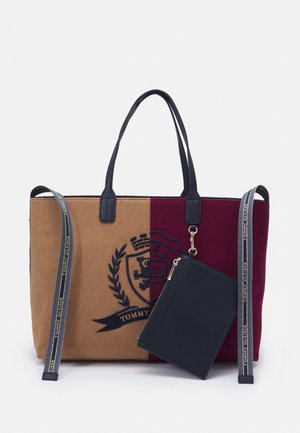 Dwukolorowa torebka typu tote w odcieniach brązu i burgundu z czarnymi uchwytami, ozdobiona herbem oraz odczepianą czarną saszetką z markowymi granatowymi paskami.