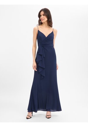 Frau trägt ein ärmelloses Abendkleid in Marineblau mit Rüschen-Detail vorne und dünnen Trägern, steht mit den Händen an den Seiten.