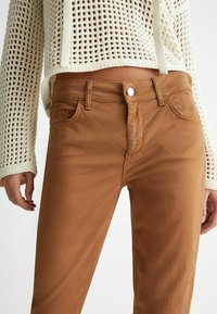 Zbliżenie na środkową część ciała w jasnobrązowych spodniach i kremowym swetrze z dzianiny szydełkowej z długimi rękawami i sznurkami.