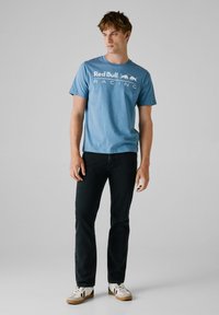 Lys blå t-shirt med korte ærmer har teksten "Red Bull Racing" i hvid. Den er parret med mørke jeans og beige sneakers med sorte detaljer.