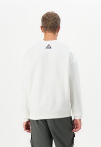 Uomo con capelli ricci corti indossa una felpa bianca con un piccolo logo triangolare sulla parte superiore della schiena e pantaloni cargo grigio scuro.