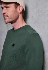 Felpa verde con texture e scollo a girocollo, dotata di un piccolo logo nero sul petto, abbinata a un cappellino beige.