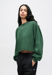 Πράσινο κοντό φούτερ με στρογγυλή λαιμόκοψη και μακριά μανίκια, με μικρό λευκό λογότυπο Nike στο στήθος, σε συνδυασμό με μαύρο παντελόνι.