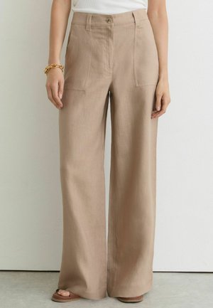 Beżowe, wysokie spodnie z szerokimi nogawkami, z przednimi kieszeniami na naszywki i zapięciem na guziki, noszone z białą bluzką, bransoletką i sandałami na szarym tle podłogi.