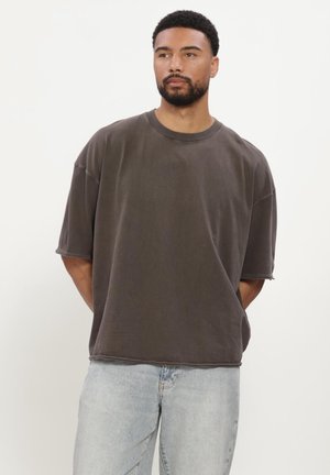 Mann mit kurzem Bart, der ein dunkles, übergroßes T-Shirt und hellblaue Jeans trägt, steht vor einfachem weißem Hintergrund.