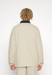 Beige jacket with a black collar, featuring long sleeves and button accents at the cuffs. Smooth texture and relaxed fit design.