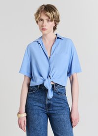 Chemise bleu clair à manches courtes avec boutons sur le devant et nouée à l'avant. Comprend une poche poitrine, fabriquée dans un tissu lisse. Portée avec un jean en denim taille haute.