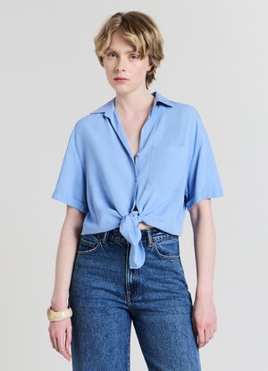 Camicia azzurro chiaro a maniche corte con bottoni e nodo frontale. Include una tasca sul petto, realizzata in tessuto liscio. Indossata con jeans di denim a vita alta.