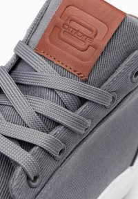 Zapatillas de lona gris con tejido texturizado, cordones redondos y un parche de cuero marrón con logo en relieve. Suela de poliuretano, diseño minimalista.