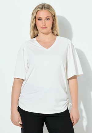 Blonde Frau mit welligem Haar trägt ein weißes V-Ausschnitt-Oberteil mit kurzen Puffärmeln und schwarze Hose, steht vor einer schlichten weißen Wand.