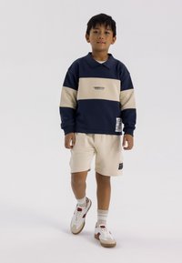 Sweatshirt bleu marine et beige à blocs de couleurs avec un col, associé à un short crème. Des sneakers blanches avec des accents rouges et beiges complètent la tenue.