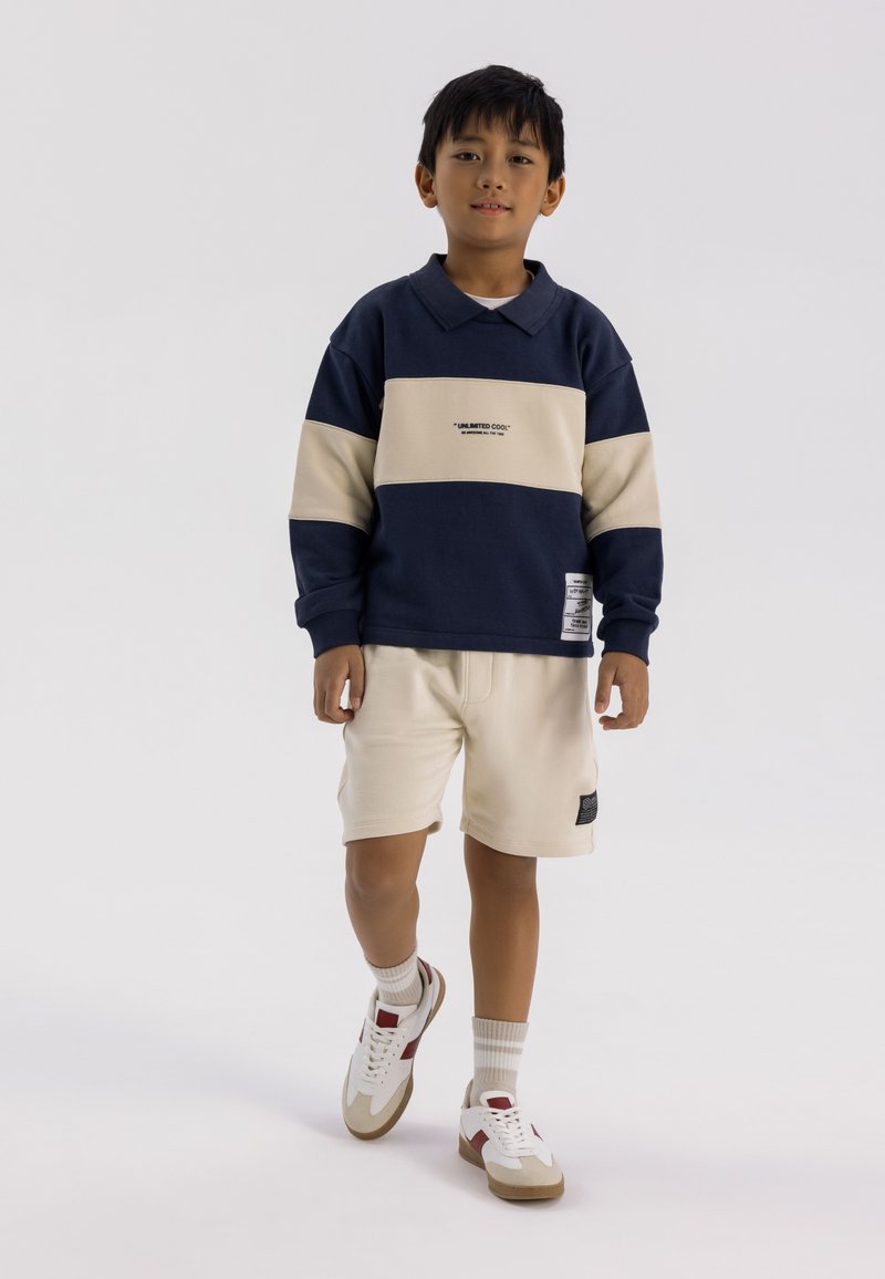 Sweatshirt bleu marine et beige à blocs de couleurs avec un col, associé à un short crème. Des sneakers blanches avec des accents rouges et beiges complètent la tenue.