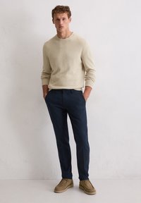 Maglione lavorato a maglia beige, pantaloni blu scuro e scarpe in suede color tan. Il maglione ha un collo rotondo e polsini a coste, mentre i pantaloni sono slim-fit.