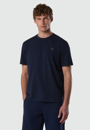 Giovane uomo con capelli ricci che indossa una maglietta a maniche corte di colore blu scuro semplice e pantaloni abbinati, in piedi davanti a uno sfondo chiaro.