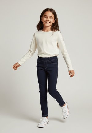 Uśmiechnięta dziewczyna z długimi brązowymi włosami, ubrana w białą bluzkę z długim rękawem, ciemne rurki i białe trampki, stojąca na jednej nodze.