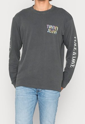 Osoba ubrana w ciemnoszarą koszulę z długim rękawem, z wielokolorowym napisem "TOMMY JEANS" na piersi oraz białym napisem "PEACE & LOVE" na prawym rękawie.