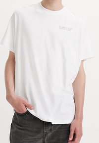 Biała bawełniana koszulka z okrągłym dekoltem, luźny krój i jasnoszary logo LEVI'S na lewej piersi. Gładka tekstura z krótkimi rękawami.