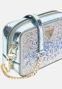 Mėlyna metalizuota rankinė su tekstūruotu paviršiumi, puošta blizgančiais rhinestones skirtingomis dydžiais, turinti auksinės spalvos grandinėlę ir logotipą.