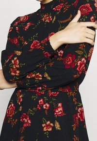 Robe noire à fleurs avec manches longues, col et poignets froncés, ornée de motifs floraux rouges et verts sur toute la surface. Tissu léger.