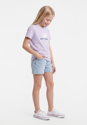 Fille souriante regardant vers le bas, main dans la poche, portant un t-shirt New Balance lavande, des shorts en jean clair et des baskets blanches.