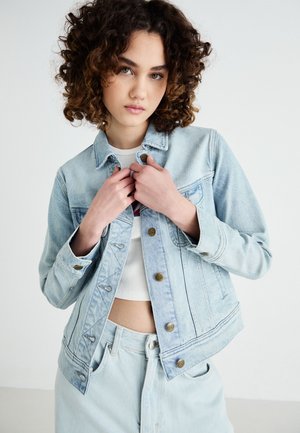 Giacca di jeans azzurro chiaro con chiusura a bottoni, due tasche sul petto, leggero sbiadimento e orlo grezzo. Indossata sopra una maglietta bianca, abbinata a pantaloni di jeans chiari.