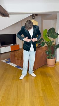 Άνδρας σε ανοιχτό τζιν και λευκά παπούτσια κουμπώνει βελούδινο σακάκι σε σκούρο χρώμα στο σαλόνι με καφέ καναπέ και μεγάλη γλάστρα φυτού.