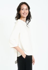 Top bianco, ampio e comodo con maniche a tre quarti, dotato di scollo rotondo e un retro leggermente più lungo, abbinato a pantaloni neri.