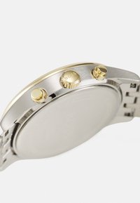 Reloj de acero inoxidable plateado con acentos dorados, caja redonda, tres botones de presión y un acabado cepillado en la correa. Fondo de caja plano.