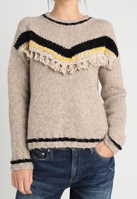 Beżowy sweter dziany z czarnymi i żółtymi paskami w kształcie zygzaków oraz frędzlami, noszony z niebieskimi dżinsami, ręka w kieszeni.