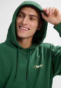 Junger Mann lächelt, trägt einen grünen Kapuzenpullover mit dem Logo "hummel SPORTSWEAR" und richtet mit der rechten Hand die Kapuze.