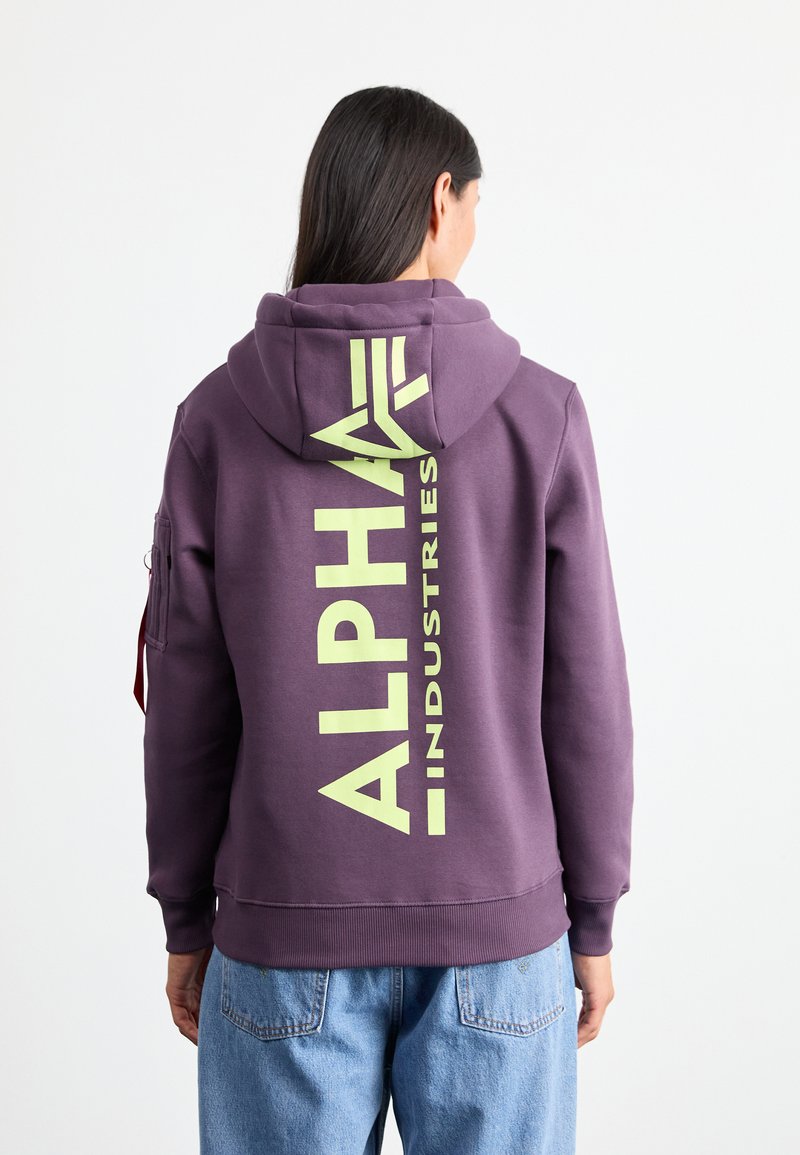 Sudadera con capucha de color púrpura oscuro con gran texto amarillo "ALPHA INDUSTRIES" en la parte trasera. Presenta un bolsillo lateral y una tela texturizada.