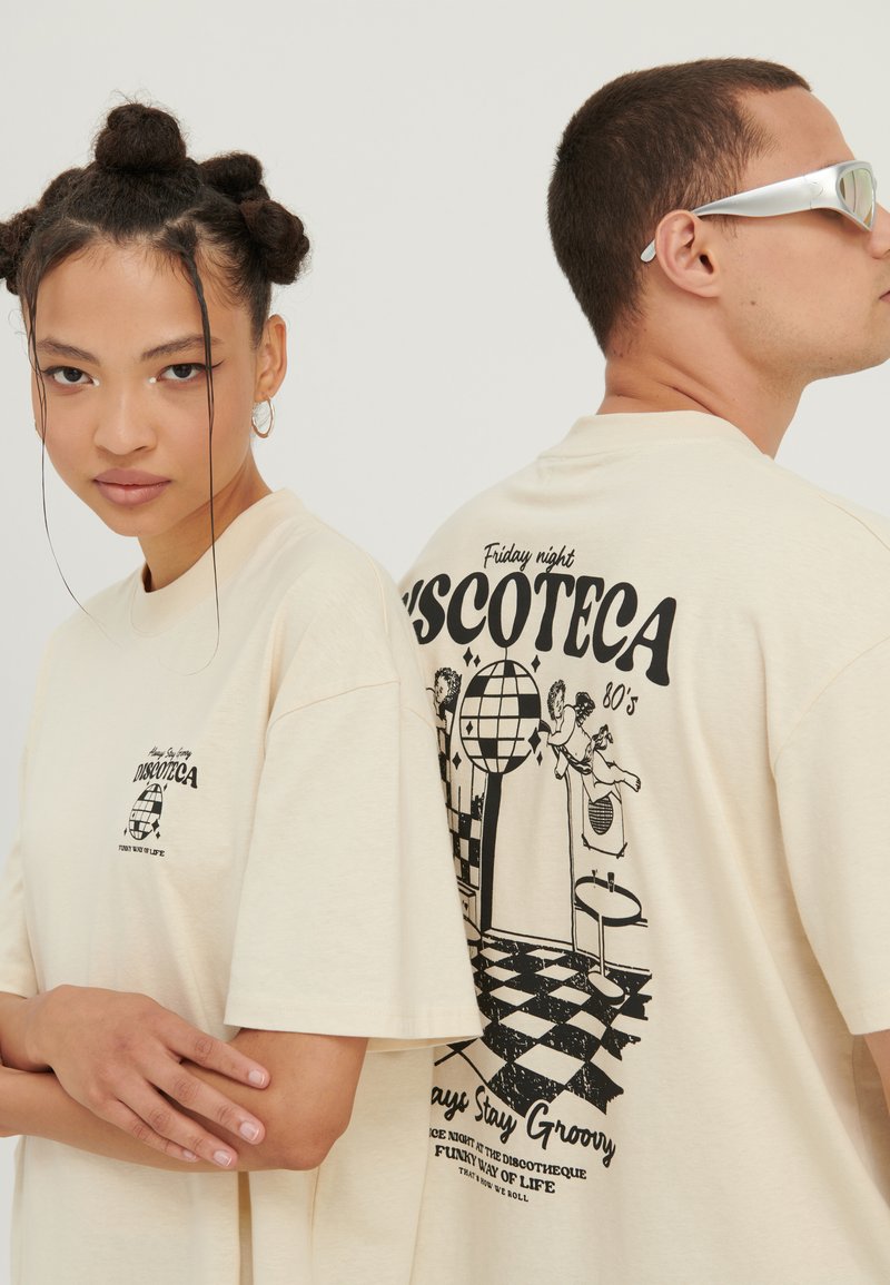Zwei Models tragen cremefarbene "Discoteca"-T-Shirts mit schwarzen, retro Disco-Motiv-Grafiken, eines zeigt die Vorderseite, das andere das Rückendesign.