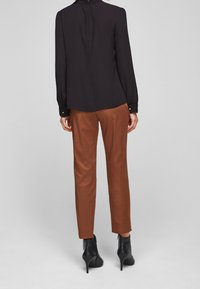 Schwarze Bluse mit langen Ärmeln, hoher Kragen und geknöpften Manschetten, kombiniert mit braunen maßgeschneiderten Hosen und schwarzen Stiefeletten.