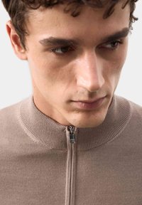 Beżowy sweter z zamkiem błyskawicznym i żeberkowym kołnierzem. Gładka tekstura, dopasowany fason i subtelne stębnowanie przy szwach.