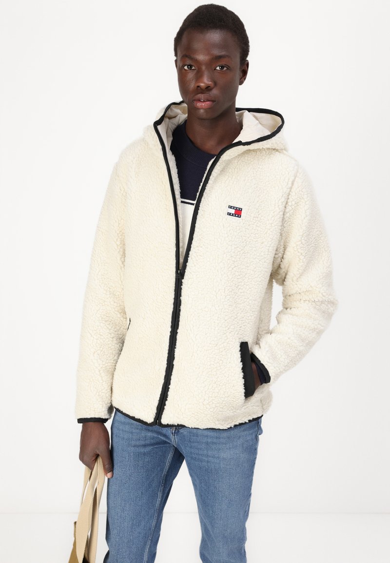 Veste en polaire crème avec fermeture éclair à l'avant, capuche et finitions noires. Elle possède des poches latérales et un petit logo sur la poitrine. Portée sur un pull bleu marine.