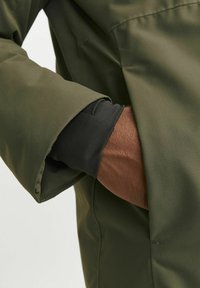 Chaqueta verde olivo con un acabado texturizado, que presenta un puño interior negro, mano parcialmente visible en el bolsillo, destacando el diseño en capas.