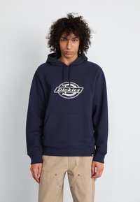 Hoodie bleu marine en tissu doux, avec un logo central en blanc. Il dispose d'une poche kangourou et de poignets côtelés pour plus de confort.