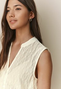 Blouse sans manches crème présentant un tissu texturé avec un motif floral subtil, un décolleté en V et des boutons décoratifs sur le devant.