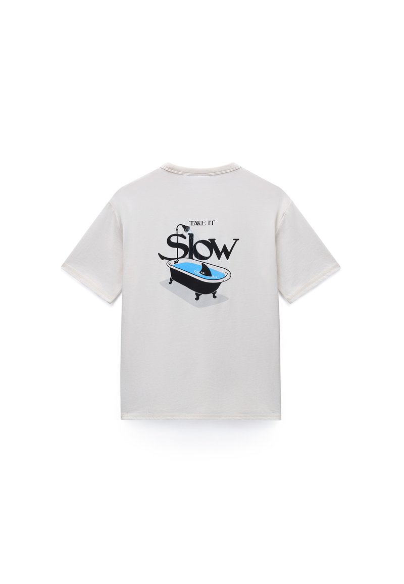 Ανοιχτό γκρι βαμβακερό t-shirt με μαύρο και μπλε γραφικό μπανιέρας και το κείμενο «Πήγαινε Αργά» στην πλάτη. Κοντά μανίκια και στρόγγυλη λαιμόκοψη.