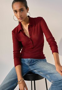 Bordeauxrotes Langarmshirt mit Kragen und Knöpfen, kombiniert mit hellblauen Jeans. Das Shirt hat einen subtilen Glanz und eine gerippte Struktur.