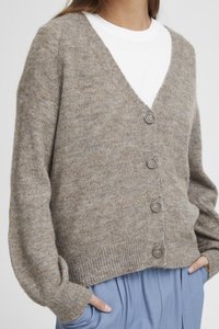 Cardigan lavorato a maglia beige con profondo scollo a V e cinque bottoni grigi. Texture morbida, vestibilità ampia e polsini a coste, indossato sopra una camicia bianca.