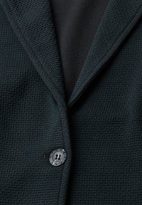 Gros plan d'un blazer texturé foncé avec un large revers et un bouton noir gravé de "STREET ONE."
