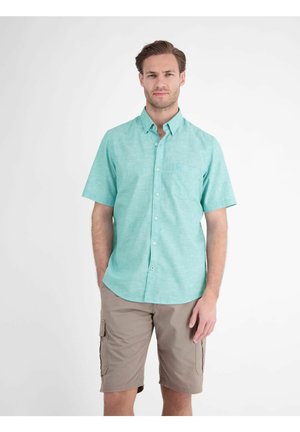Man staat met een lichtblauw overhemd met korte mouwen en beige cargoshorts tegen een effen witte achtergrond.