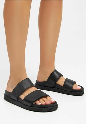 Sandales à glissière en cuir noir avec deux larges brides, semelle plate et design à bout ouvert. Détail de couture sur la bride supérieure.